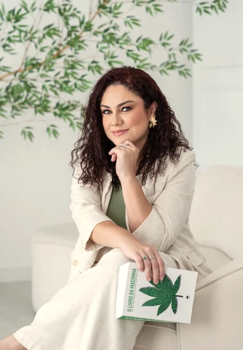 Retrato da Dra. Yasmin Becker sentada em poltrona clara, com O Livro da Maconha nas mãos e galhos de planta ao fundo