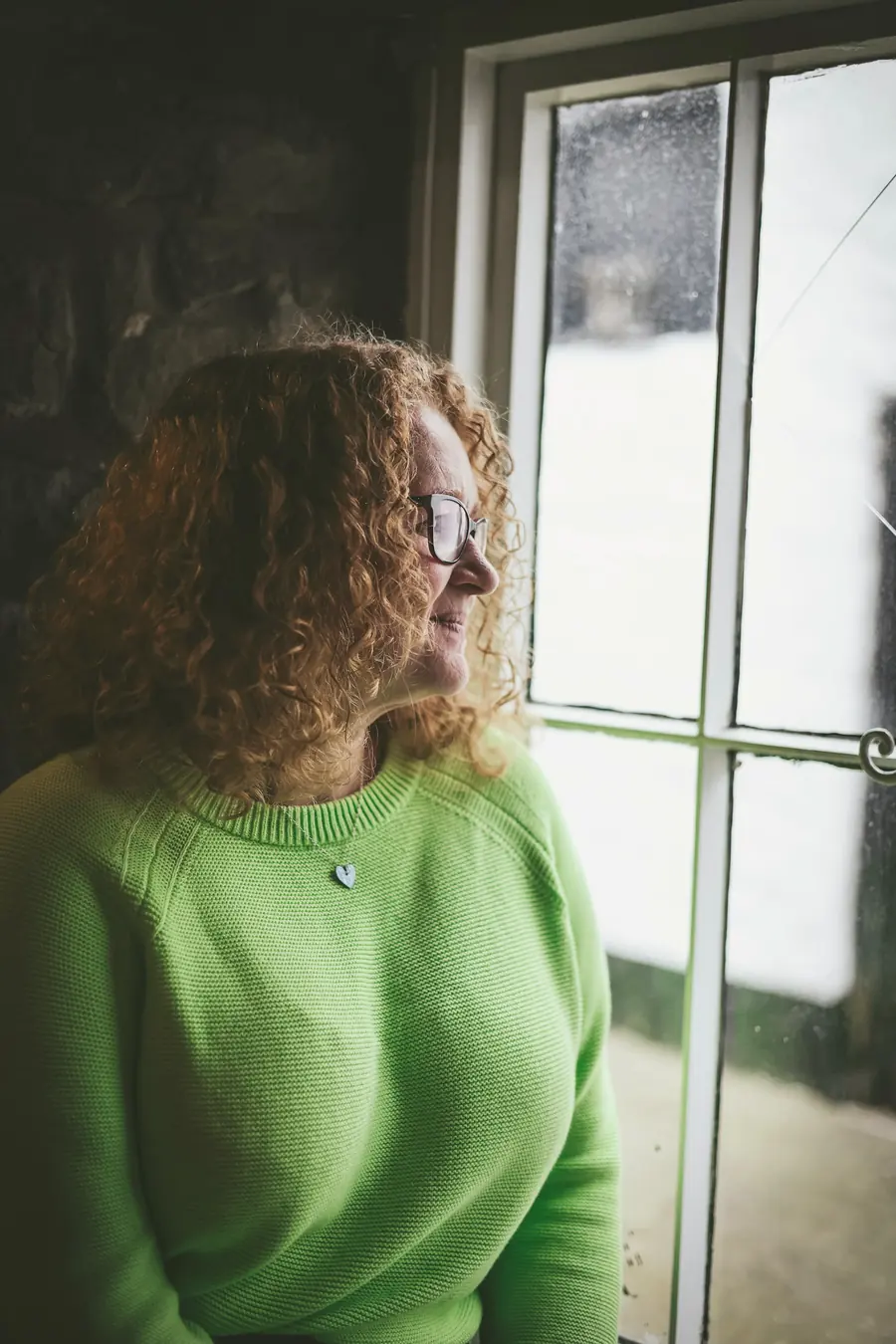 Mulher de suéter verde olhando pela janela, pensativa