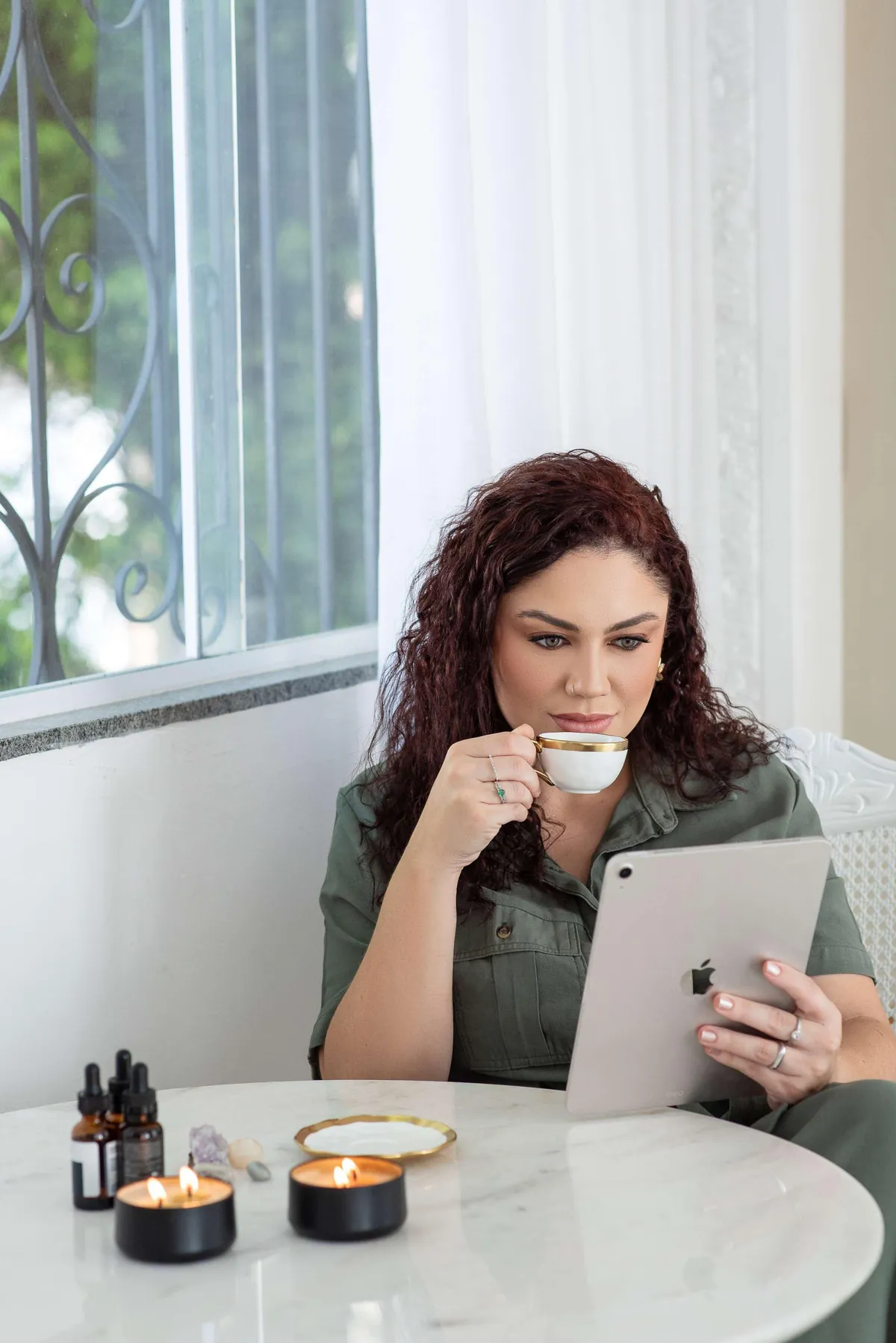 Dra. Yasmin Becker lendo num tablet, com uma xícara na mão, numa mesa com frascos conta-gotas e velas